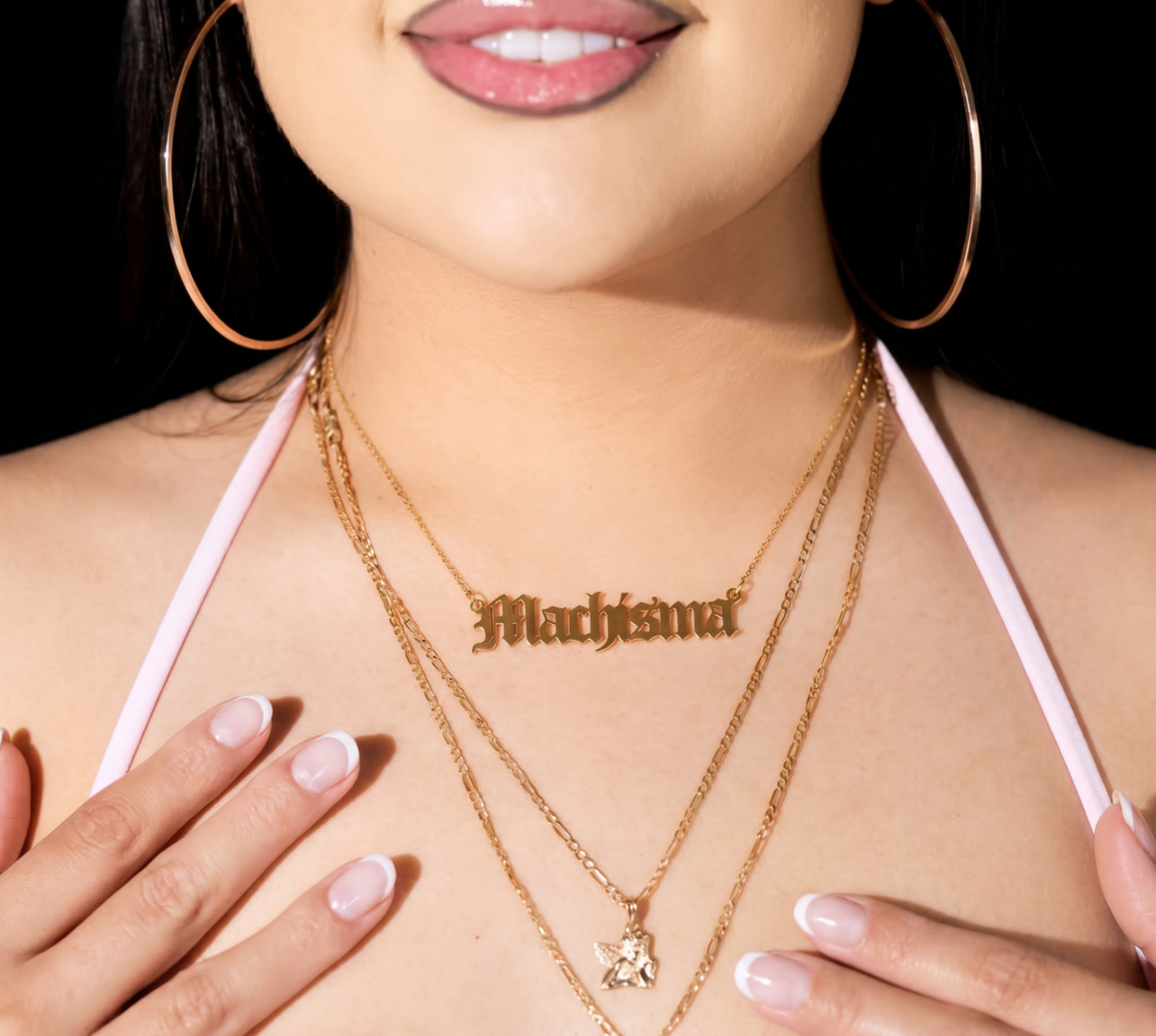 Close-up of a woman wearing a gold 'Machisma' nameplate necklace with layered gold chains and large hoop earrings
